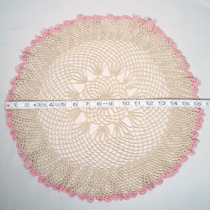 May include: A beige and pink crocheted doily, approximately 16 inches in diameter. The doily features a central floral design and a scalloped pink border. The intricate openwork pattern is visible throughout the piece.