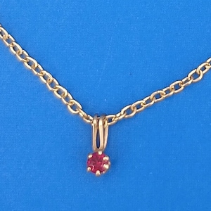 May include: A gold-coloured necklace featuring a small, round, red gemstone pendant. The chain has a simple link design, and the pendant is held by a gold-coloured setting. The background is a solid blue.