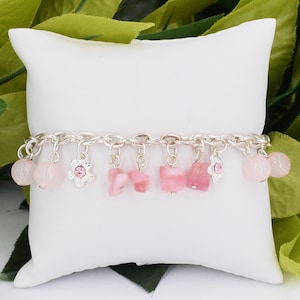 May include: A silver charm bracelet with pink stone and flower charms. The bracelet is on a white pillow.