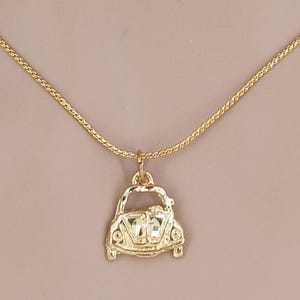 May include: Gold-colored necklace featuring a pendant shaped like a vintage car. The pendant has detailed features, including headlights and a front grill. The chain is a delicate, textured design. The necklace is displayed against a neutral background.