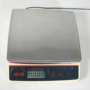 May include: A rectangular electronic scale with a large, flat, brushed stainless steel weighing platform. The base is off-white with a black control panel featuring the red text MARS, a digital display, and buttons labelled ON TARE, MODE, and OFF.