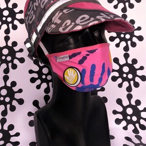 May include: Pink face mask with a colourful design featuring handprints and a yellow circle with a hand symbol. The mask has the word "Deemboy" printed on it. The mask is worn by a mannequin wearing a pink and grey cap with a reflective strip.