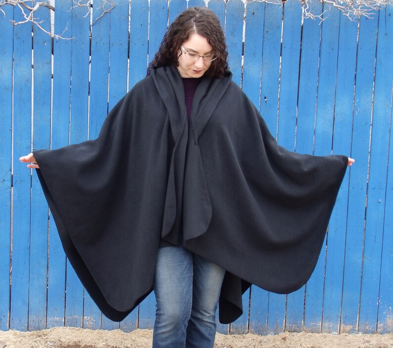 Op de afbeelding: Een zwarte fleece cape met een capuchon, gedragen over een spijkerbroek. De cape is over de schouders gedrapeerd en heeft een losse, vloeiende pasvorm.