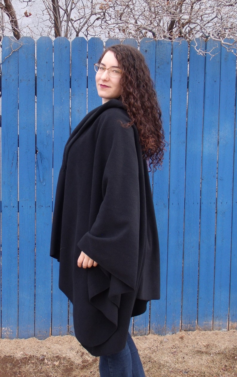 Op de afbeelding: Een zwarte fleece-wrap met een gedrapeerd ontwerp. De wrap wordt gedragen door een persoon met krullend bruin haar en een bril, die voor een blauwe houten schutting staat.
