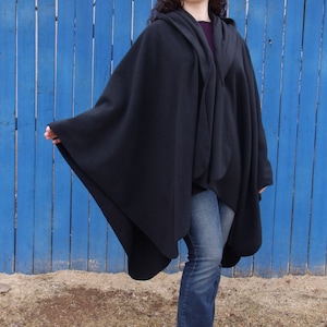 Op de afbeelding: Een zwarte capuchon met een lang, vloeiend ontwerp. De cape wordt gedragen over een blauwe jeans en de persoon draagt zwarte schoenen.
