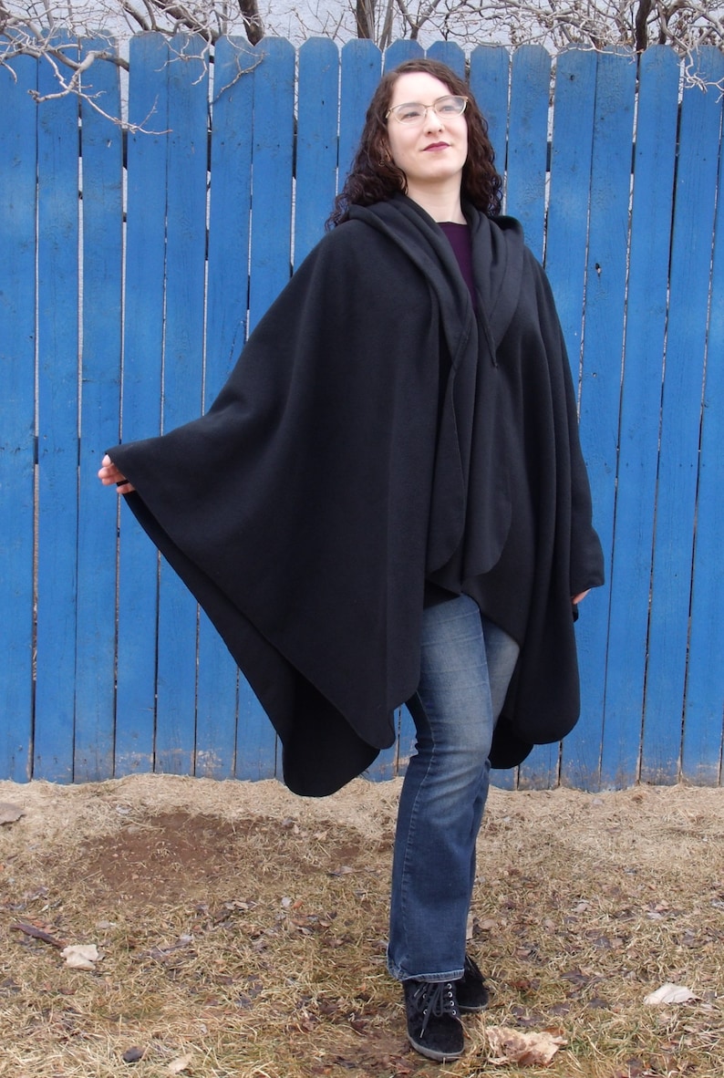 Op de afbeelding: Een zwarte capuchon met een lang, vloeiend ontwerp. De cape wordt gedragen over een blauwe jeans en de persoon draagt zwarte schoenen.