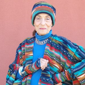 May include: A woman wearing a blue turtleneck sweater and a colorful patterned fleece poncho with matching fingerless gloves. She is also wearing a colorful patterned fleece hat.
