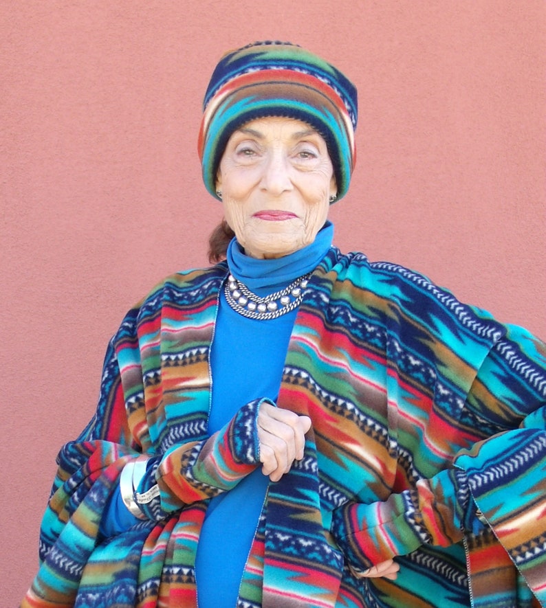 May include: A woman wearing a blue turtleneck sweater and a colorful patterned fleece poncho with matching fingerless gloves. She is also wearing a colorful patterned fleece hat.