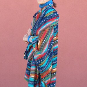 May include: A woman wearing a colorful patterned poncho with a blue turtleneck sweater underneath. The poncho has a geometric design with shades of blue, red, orange, and brown.