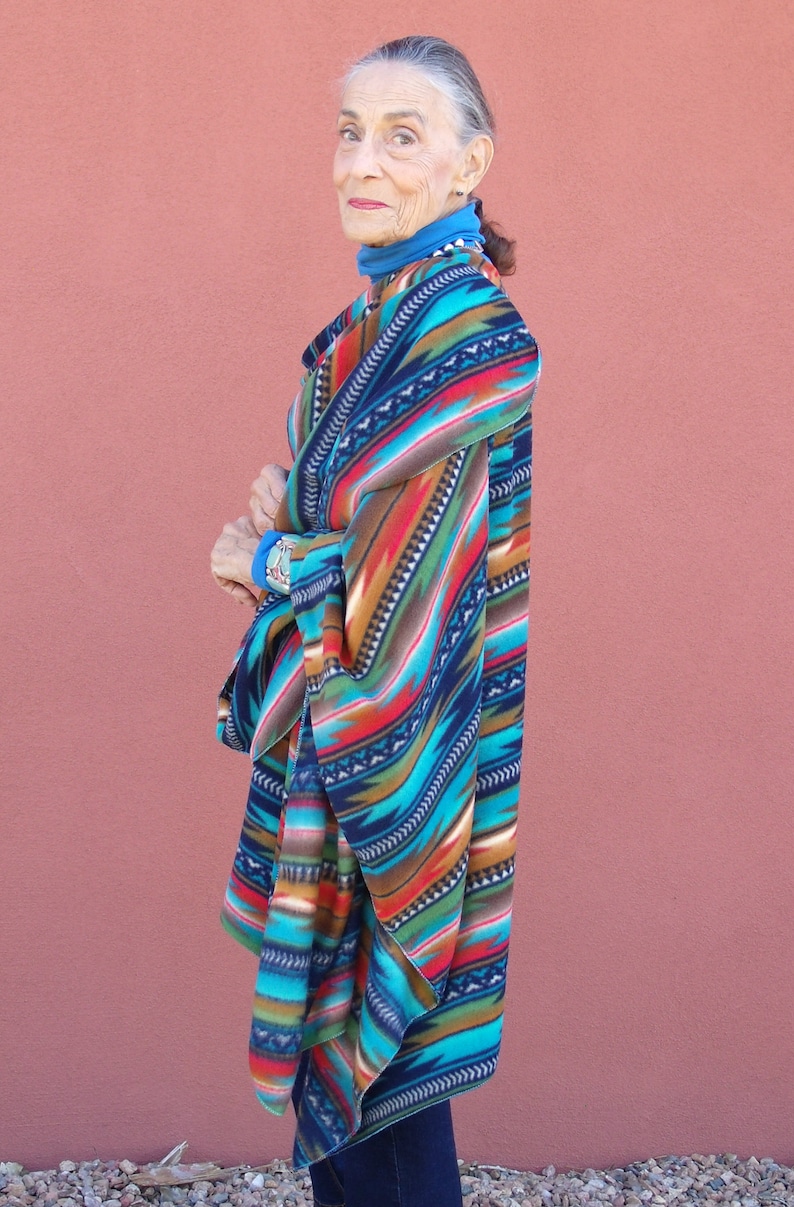 May include: A woman wearing a colorful patterned poncho with a blue turtleneck sweater underneath. The poncho has a geometric design with shades of blue, red, orange, and brown.