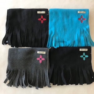 May include: Four fleece scarves with fringe. Each scarf features a different color: black, blue, gray, and dark blue. Each scarf has a small tag with the text "Fleece Studio" and a stylized symbol of a sun with four rays.