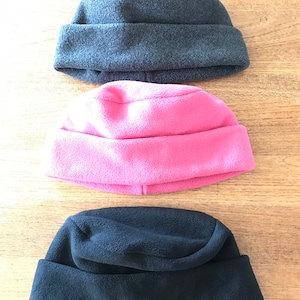 May include: Three fleece hats in different colors: gray, pink, and black. The hats are folded in half and placed on a wooden surface.