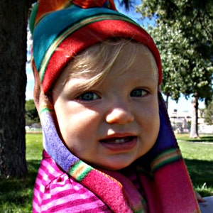 May include: A young child wearing a colorful striped hat and matching scarf. The hat has a pointed top and the scarf is tied around the child's neck. The child is wearing a pink and white striped shirt.