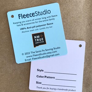 May include: A light blue tag with the text "FleeceStudio" and information about the company's fleece products. The tag also includes a logo for "NM True Certified". The back of the tag is white with a blank space for "Style", "Color/Pattern", and "Size". The text "Thank you for buying a handmade product" is at the bottom of the tag.