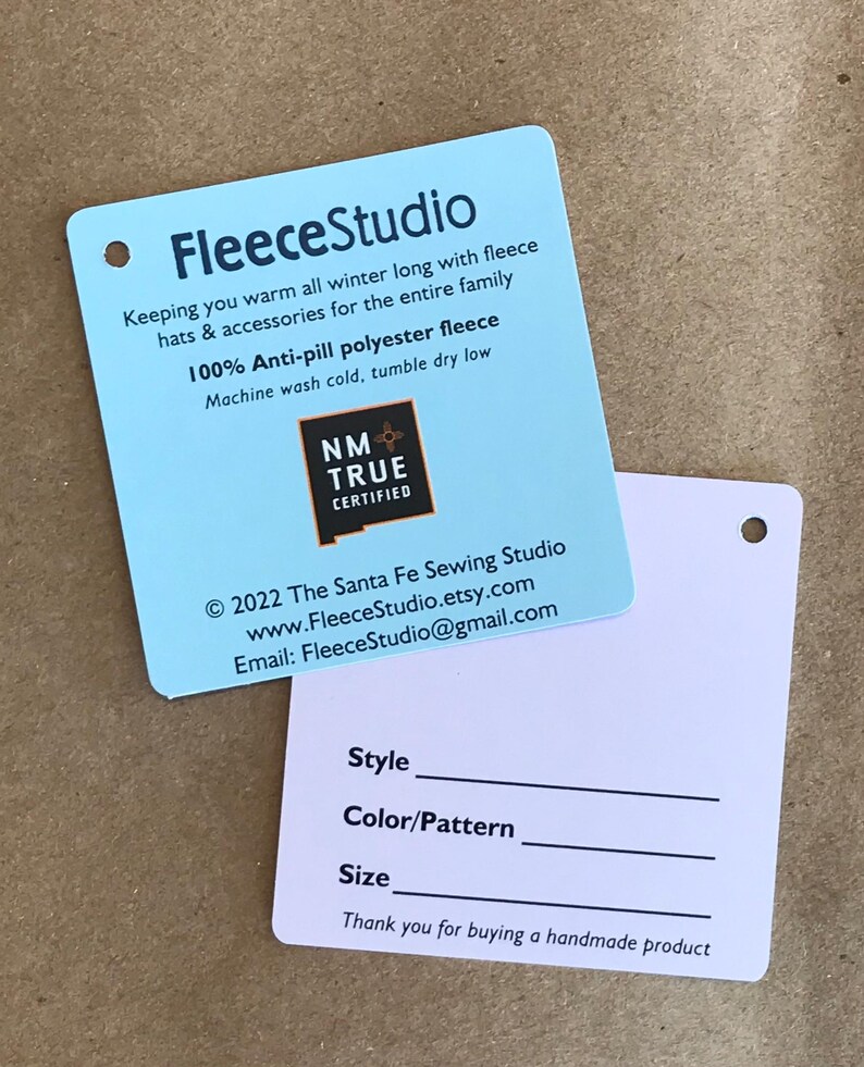 May include: A light blue tag with the text "FleeceStudio" and information about the company's fleece products. The tag also includes a logo for "NM True Certified". The back of the tag is white with a blank space for "Style", "Color/Pattern", and "Size". The text "Thank you for buying a handmade product" is at the bottom of the tag.