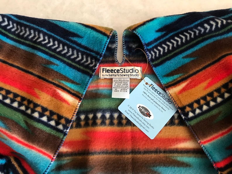 May include: A multi-colored fleece blanket with a Southwestern pattern. The blanket is folded in half and has a tag attached that reads "Fleece Studio by the Santa Fe Sewing Studio".
