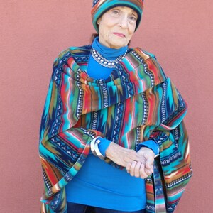 May include: A woman wearing a blue turtleneck sweater, blue jeans, and a colorful patterned poncho. She is also wearing a colorful patterned hat and a silver necklace.