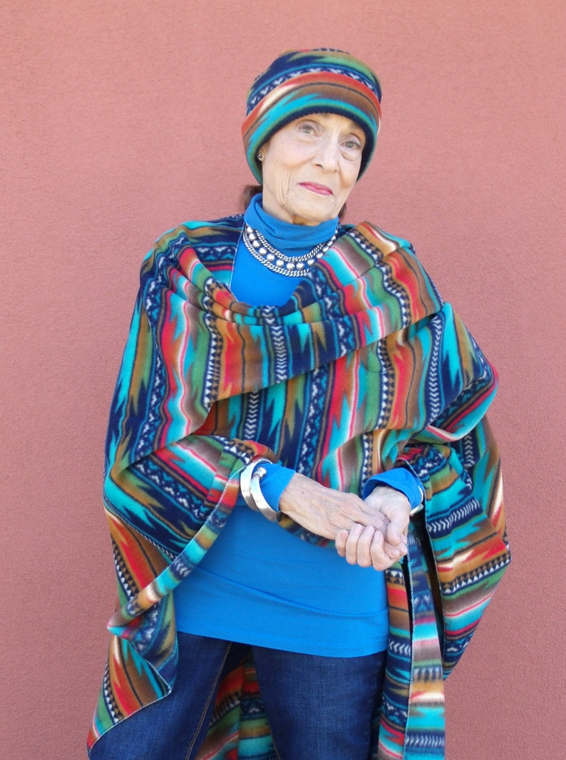 May include: A woman wearing a blue turtleneck sweater, blue jeans, and a colorful patterned poncho. She is also wearing a colorful patterned hat and a silver necklace.