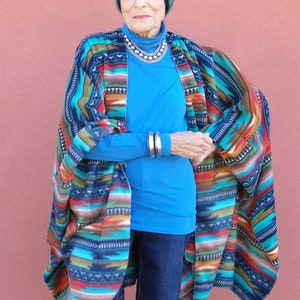 May include: A woman wearing a blue shirt and jeans with a colorful patterned poncho and a knitted hat.