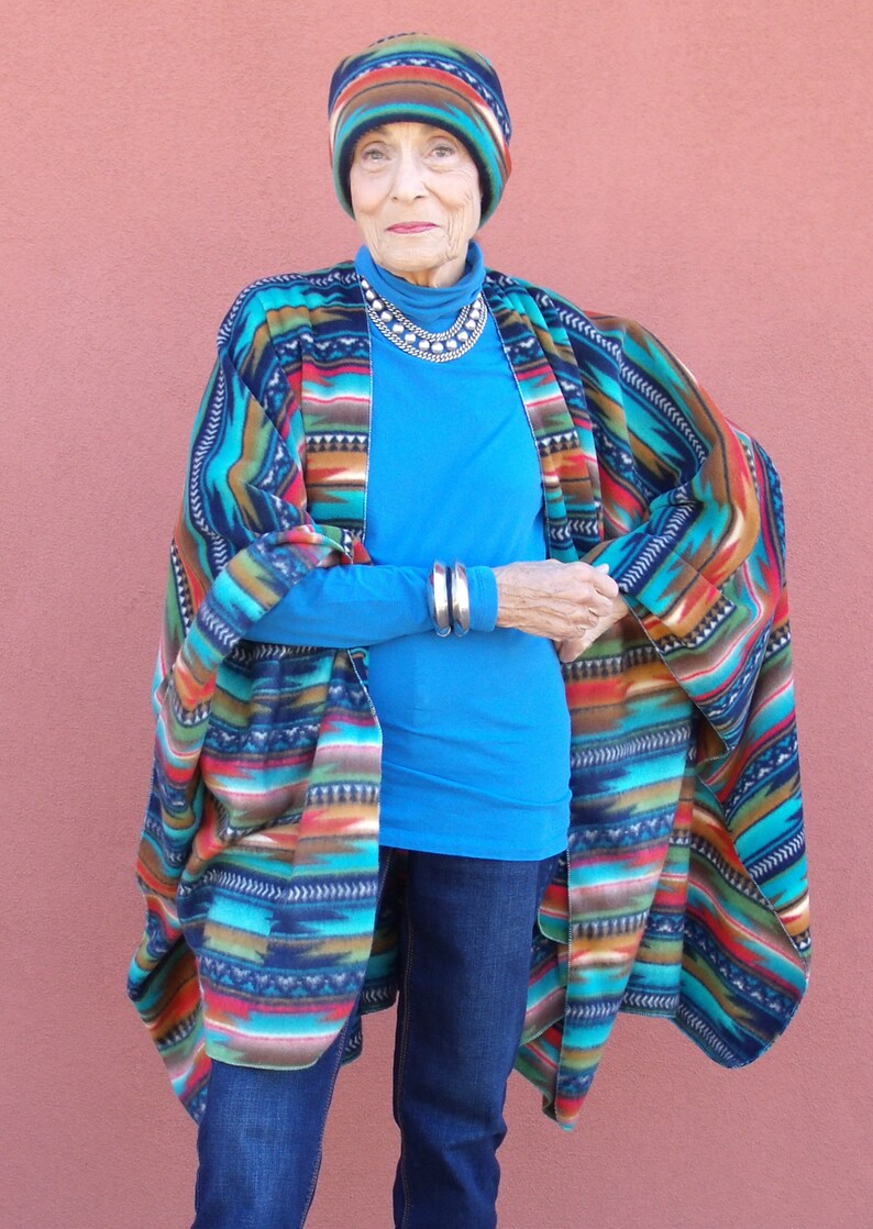 May include: A woman wearing a blue shirt and jeans with a colorful patterned poncho and a knitted hat.