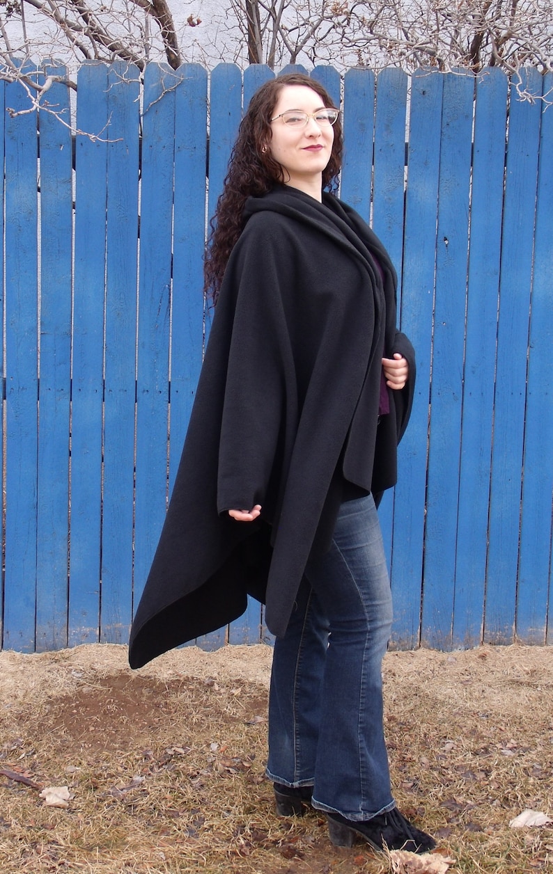 Op de afbeelding: Een vrouw draagt een zwarte cape met een lang, vloeiend ontwerp. Ze staat voor een blauwe houten schutting en draagt een blauwe jeans en zwarte schoenen.