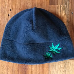 May include: A dark blue fleece beanie with a green embroidered marijuana leaf design.