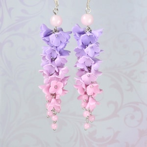 Peut inclure: Une paire de boucles d'oreilles pendantes avec un d&eacute;grad&eacute; de fleurs en p&acirc;te polym&egrave;re violettes et roses. Les boucles d'oreilles sont faites avec des crochets et des perles argent&eacute;s.