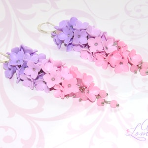 Peut inclure: Une paire de boucles d'oreilles pendantes avec un d&eacute;grad&eacute; de fleurs violettes et roses. Les fleurs sont en p&acirc;te polym&egrave;re et sont enfil&eacute;es sur un fil d'argent. Les boucles d'oreilles sont finies avec de petites perles argent&eacute;es.