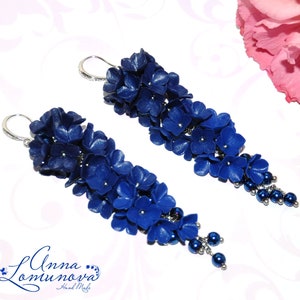 May include: A pair of long, dangling earrings featuring clusters of deep blue flowers. The earrings have silver-colored hooks and are accented with small, dark blue beads. A pink flower and a yellow-green flower are in the background.