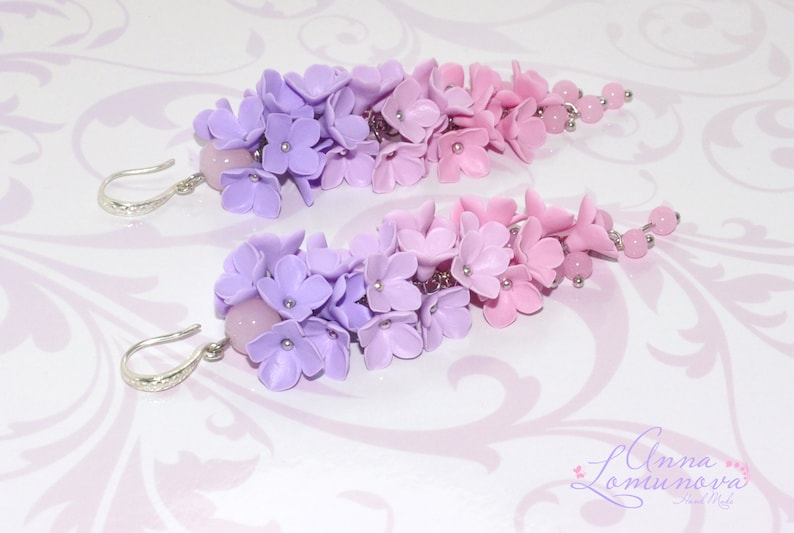 Peut inclure: Une paire de boucles d'oreilles pendantes avec un d&eacute;grad&eacute; de fleurs en p&acirc;te polym&egrave;re violettes et roses. Les boucles d'oreilles sont faites avec des crochets argent&eacute;s et ont une perle rose en haut.