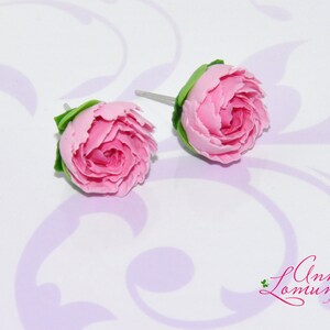 Pode incluir: Um par de brincos de argila polim&eacute;rica rosa em forma de pe&ocirc;nias. Os brincos est&atilde;o sobre um fundo branco com um padr&atilde;o floral roxo claro.