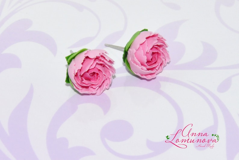 Pode incluir: Um par de brincos de argila polim&eacute;rica rosa em forma de pe&ocirc;nias. Os brincos est&atilde;o sobre um fundo branco com um padr&atilde;o floral roxo claro.