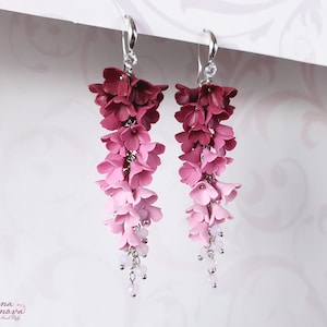 May include: A pair of dangle earrings with a gradient of pink and red polymer clay flowers. The earrings have silver hooks and a small crystal charm at the bottom.