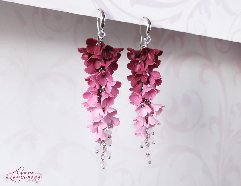 May include: A pair of dangle earrings with a gradient of pink and red polymer clay flowers. The earrings have silver hooks and a small crystal charm at the bottom.