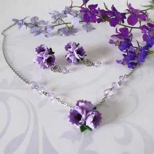 May include: A delicate silver necklace with a purple flower pendant and matching earrings. The necklace features small crystal accents and the flower pendant is made of polymer clay.