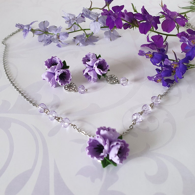 May include: A delicate silver necklace with a purple flower pendant and matching earrings. The necklace features small crystal accents and the flower pendant is made of polymer clay.