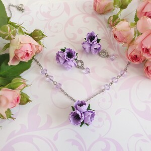 May include: A delicate silver necklace and earrings set with a cluster of three purple flowers. The necklace features a silver chain with clear crystal beads and the earrings have a single crystal dangle.