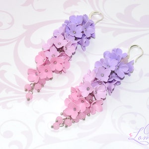 Peut inclure: Une paire de boucles d'oreilles pendantes avec un d&eacute;grad&eacute; de fleurs roses et violettes. Les boucles d'oreilles sont en p&acirc;te polym&egrave;re et ont des crochets argent&eacute;s.