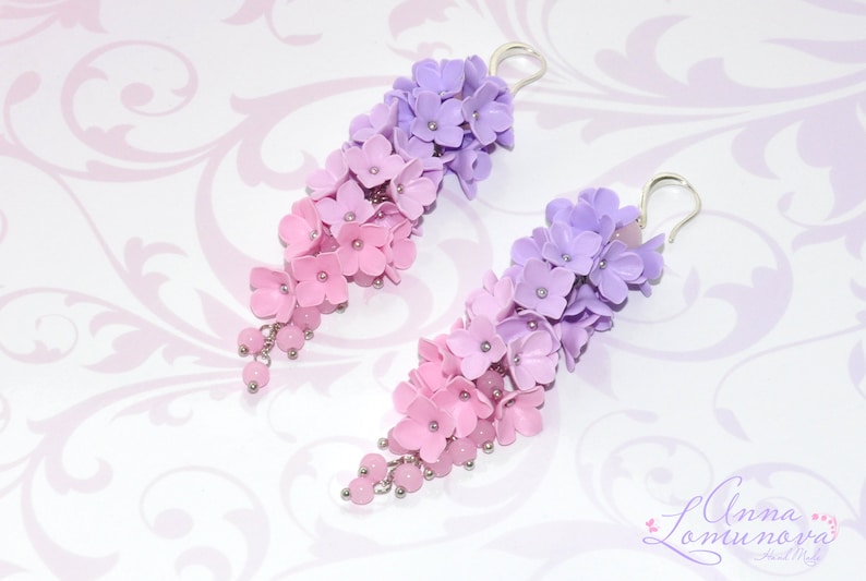 Peut inclure: Une paire de boucles d'oreilles pendantes avec un d&eacute;grad&eacute; de fleurs roses et violettes. Les boucles d'oreilles sont en p&acirc;te polym&egrave;re et ont des crochets argent&eacute;s.