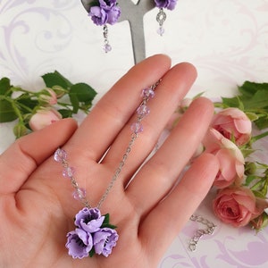 May include: A delicate necklace and earrings set featuring a cluster of three lavender polymer clay flowers. The necklace has a silver chain and clear crystal beads. The earrings are silver with a single lavender flower.