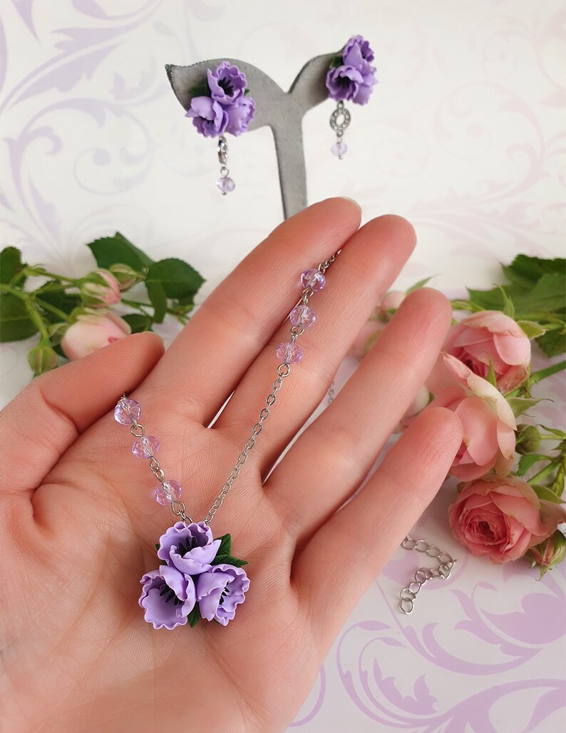 May include: A delicate necklace and earrings set featuring a cluster of three lavender polymer clay flowers. The necklace has a silver chain and clear crystal beads. The earrings are silver with a single lavender flower.