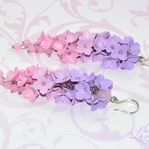 Peut inclure: Une paire de boucles d'oreilles pendantes avec un d&eacute;grad&eacute; de fleurs en p&acirc;te polym&egrave;re rose et violette. Les boucles d'oreilles sont faites avec des crochets en m&eacute;tal argent&eacute;.