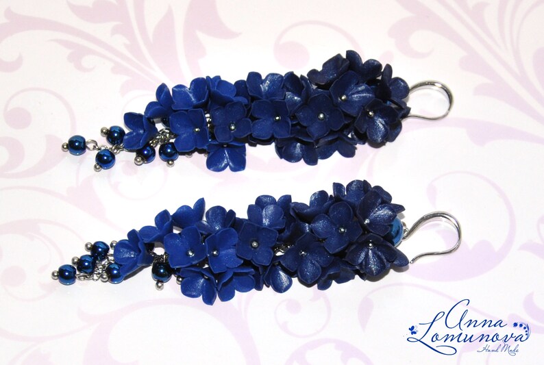 May include: A pair of dark blue floral earrings. Each earring features clusters of small, dark blue flowers with silver accents and dangling dark blue beads. The earrings have silver-colored ear wires.