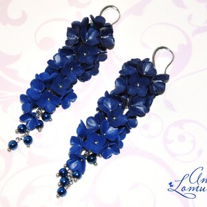 May include: A pair of long, dangling earrings featuring clusters of deep blue flowers and small, dark blue beads. The floral design is accented with silver-colored metal findings. The earrings are set against a soft, patterned background.