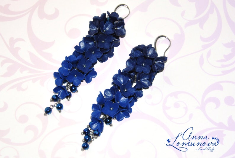 May include: A pair of long, dangling earrings featuring clusters of deep blue flowers and small, dark blue beads. The floral design is accented with silver-colored metal findings. The earrings are set against a soft, patterned background.