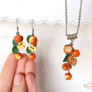 May include: A set of earrings and a necklace featuring a cluster of red and yellow apple charms with green leaves. The charms are made of polymer clay and resemble cut apples with visible seeds.