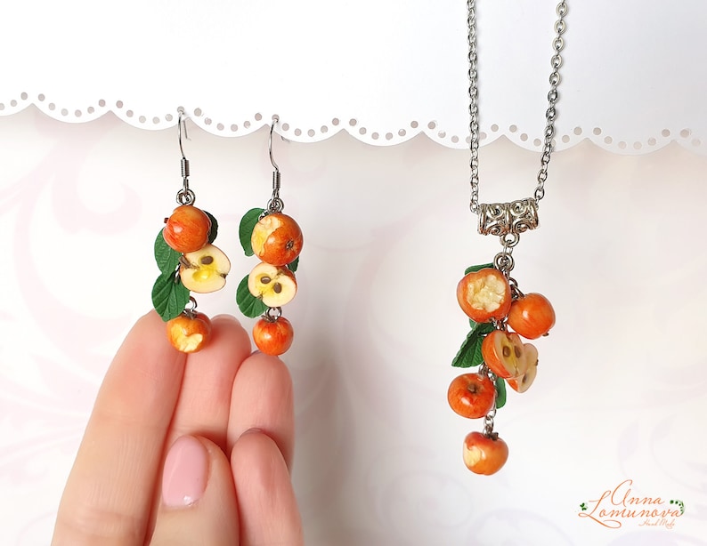 May include: A set of earrings and a necklace featuring a cluster of red and yellow apple charms with green leaves. The charms are made of polymer clay and resemble cut apples with visible seeds.