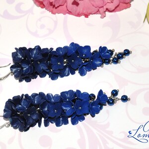 May include: A pair of handcrafted, dark blue floral earrings. Each earring features a cluster of small, flower-shaped elements and a beaded accent. The earrings have silver-colored ear wires and are set against a floral background.