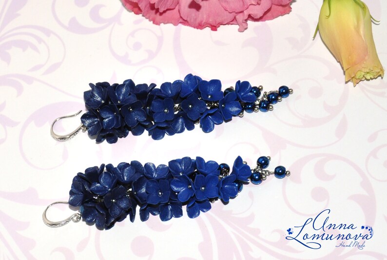 May include: A pair of handcrafted, dark blue floral earrings. Each earring features a cluster of small, flower-shaped elements and a beaded accent. The earrings have silver-colored ear wires and are set against a floral background.