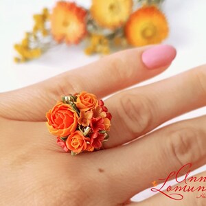 Peut inclure: Une bague orn&eacute;e d'un bouquet de fleurs en p&acirc;te polym&egrave;re orange et rouge. La bague est port&eacute;e au doigt.